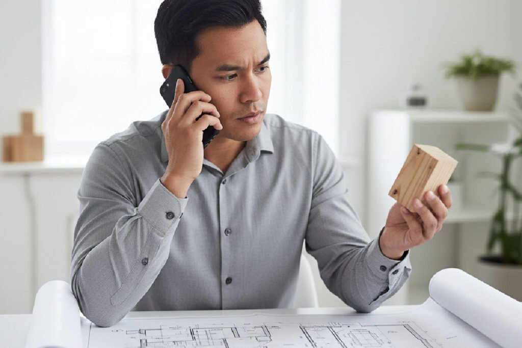 A designer on the phone looking concerned at a wood sample, illustrating the challenge of sourcing from an unreliable custom furniture manufacturer for a bespoke furniture project.