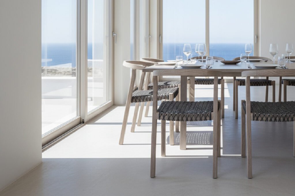 A minimalist sungkai wood dining chair in dining room area with Mediterranean style furniture, overlooking a bright blue ocean. A long, light wood dining table is set with glassware, surrounded by modern woven rattan chairs. This image represents commercial-grade furniture suitable for a luxury coastal resort.