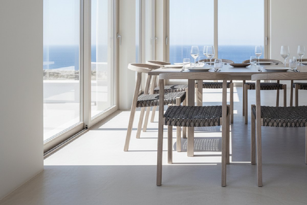 A minimalist sungkai wood dining chair in dining room area with Mediterranean style furniture, overlooking a bright blue ocean. A long, light wood dining table is set with glassware, surrounded by modern woven rattan chairs. This image represents commercial-grade furniture suitable for a luxury coastal resort.