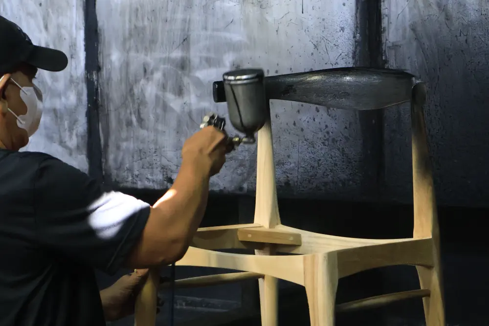 A technician in a dedicated Spray Booth applying finish to a chair, ensuring a dust-free and even coating at an Indonesian furniture manufacturer.