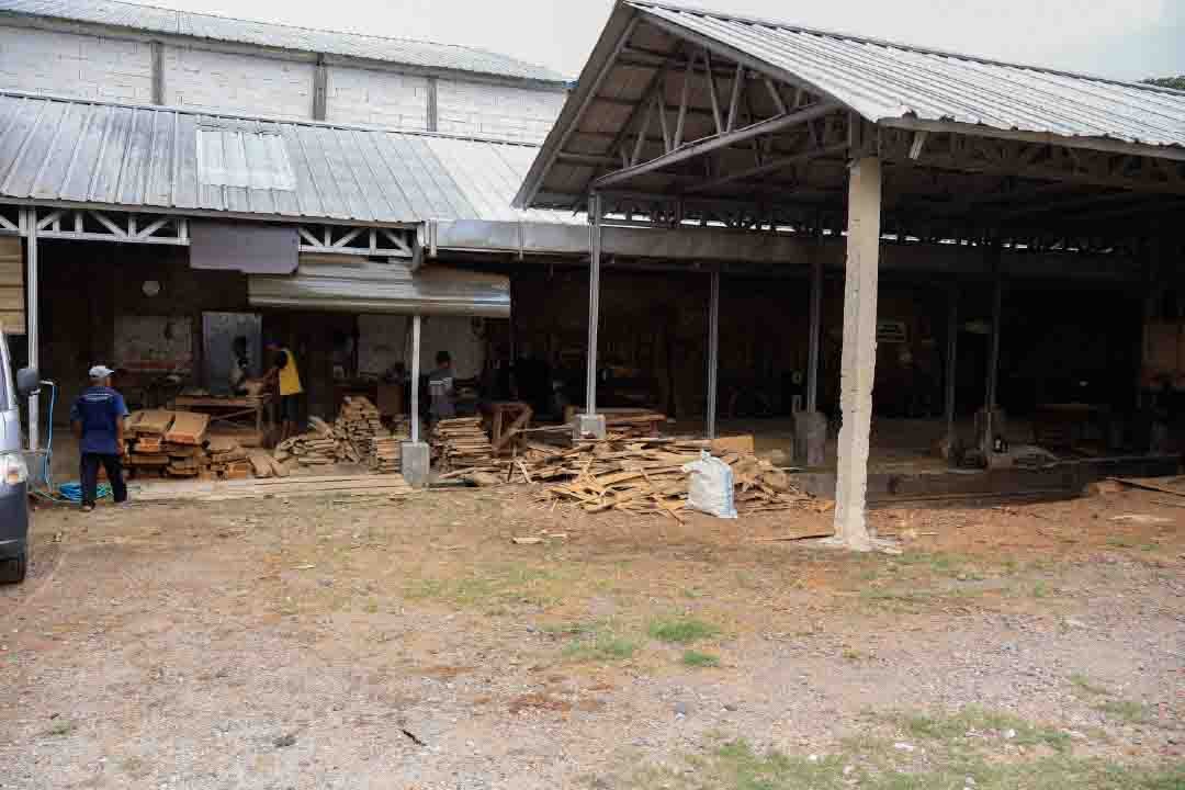 Exterior of a large-scale Indonesian furniture factory facility with a corrugated metal roof and open-air timber processing areas. The foreground displays piles of raw wood scraps and lumber ready to be processed into export-quality furniture components.