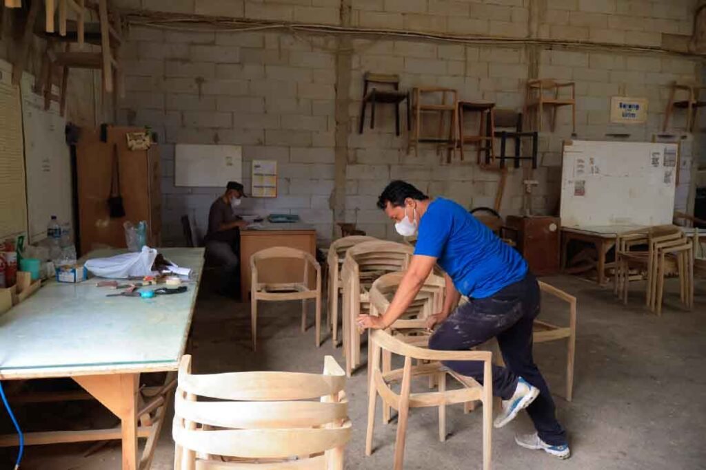 Interior view of an active Indonesian furniture manufacturing workshop, featuring a craftsman in a blue shirt inspecting a sanded wooden chair frame. The background shows a busy production floor with stacked chair components, hanging frames, and administrative staff managing orders.