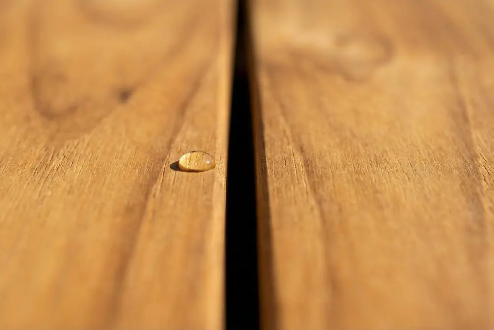 Macro close-up of a weather-resistant outdoor table surface, highlighting the durability of Indonesian furniture. The image focuses on a precise gap between golden teak planks where a single water droplet rests, demonstrating the wood's natural resistance to moisture.