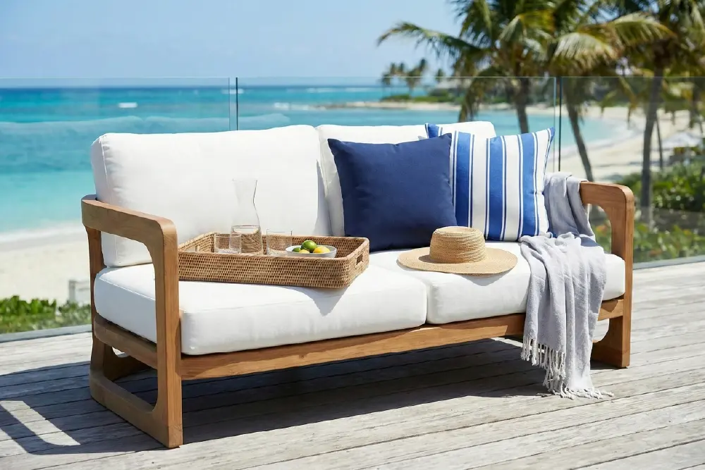 Page Optimize_3-01 MPP Furniture outdoor teak two-seater sofa featuring plush white weather-resistant cushions and nautical blue striped throw pillows, styled on a sun-bleached wooden deck with a turquoise ocean background to showcase luxury coastal living.