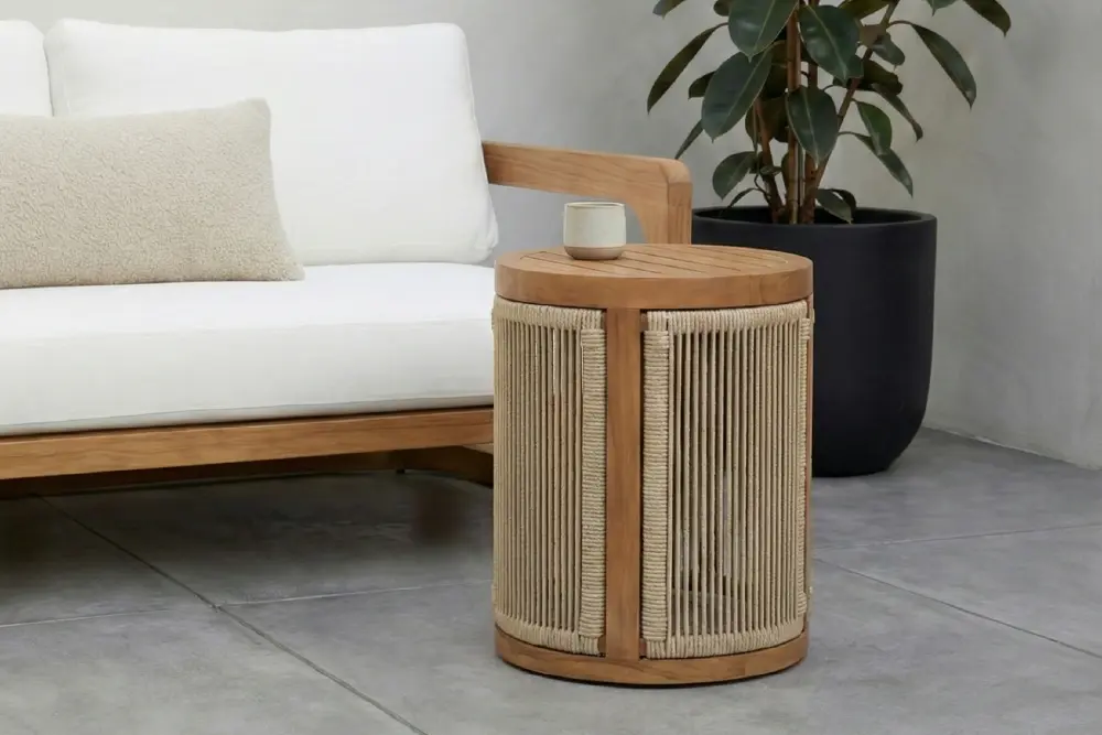 Page Optimize_3-02 Close-up of an MPP Furniture round teak side table featuring intricate vertical beige rope weaving and a slatted wood top, displayed in a modern minimalist interior next to a white sofa and a potted rubber plant.