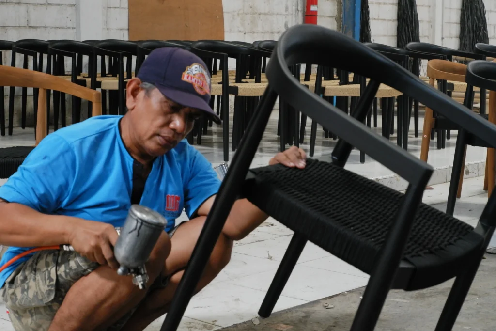 An Indonesian craftsman applying a protective spray finish to a woven chair, ensuring high-quality durable furniture for retailer costs reduction by preventing premature wear in retail environments.