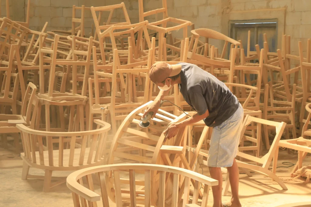 Craftsman carefully sanding a curved wooden chair frame in a factory, a crucial preparation step that determines how material affect furniture lifespan and durability.