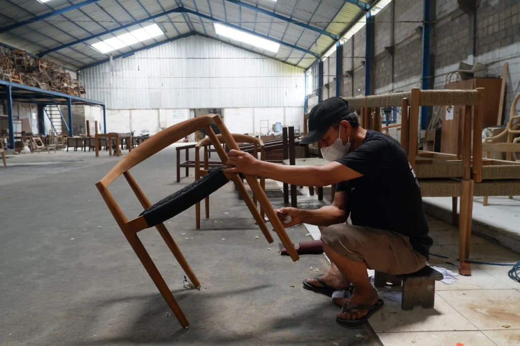 A skilled craftsman meticulously finishing a wooden chair in a large facility, showcasing the manufacturing capability and quality control behind Indonesia custom furniture sourcing.