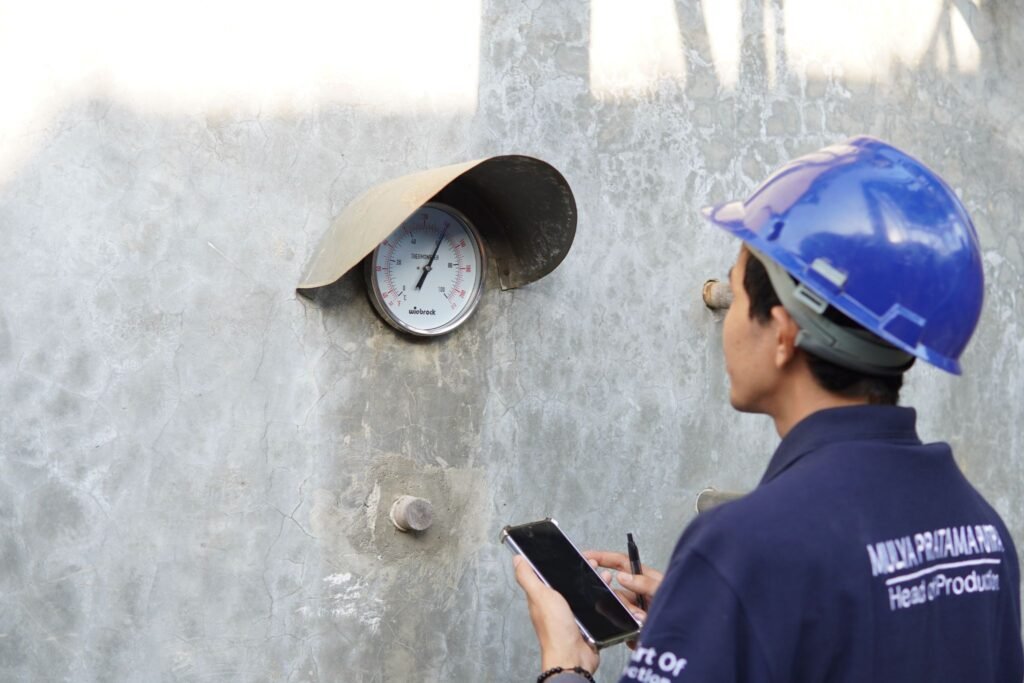Indonesian furniture worker monitoring kiln drying temperature and recording production data in a furniture manufacturing facility.